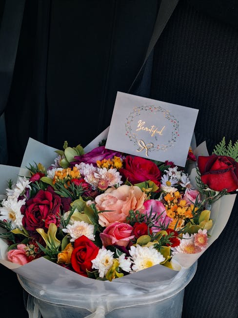 A vibrant floral bouquet arranged with a variety of fresh flowers including deep red roses, pink roses, peach-colored roses, white daisies, and small yellow and orange blooms, all set within white wra