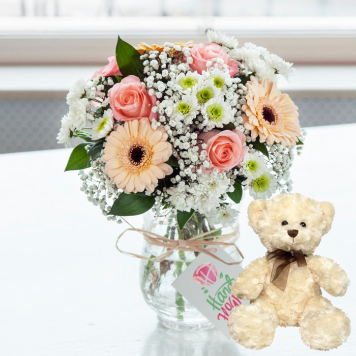 On a flawlessly white, gently reflective table, a delicate anniversary scene is composed with the kind of understated charm that suits the leafy streets around Crystal Palace. At the centre of the image, a clear glass jar-style vase displays a rounded, carefully balanced bouquet. The flower mix leans into soft pastels: blush pink roses with velvety petals, their edges just curling outward, and peach gerbera daisies featuring velvety dark brown centres that anchor the design. Around these focal blooms, white chrysanthemums accented with hints of lime green create a scalloped ring of texture, while threads of white baby's breath drift between them, filling any gaps with an airy, cloudlike feel. The stems are visible through the glass, adding a sense of freshness and honesty to the arrangement. Around the neck of the vase, a slender band of natural twine is tied and finished with a simple bow, holding a small white rectangular tag. A pink heart icon and the word Handmade written in green, cursive letters quietly indicate a handcrafted creation, as if assembled by an attentive local florist. To the right, a cream teddy bear sits upright, leaning ever so slightly toward the flowers. Its fur looks softly textured, almost woolly, with shiny dark bead eyes that catch the light, a brown button nose, and a slim satin ribbon tied around its neck. The background is bright and out of focus, suggesting daylight streaming through a nearby window, while keeping all attention on the tender pairing. The combination of pastel bouquet and cuddly bear evokes warmth, reassurance, and celebration, ideal for marking anniversaries or heartfelt milestones in Crystal Palace, from the triangle's cafés up to the quieter residential roads.